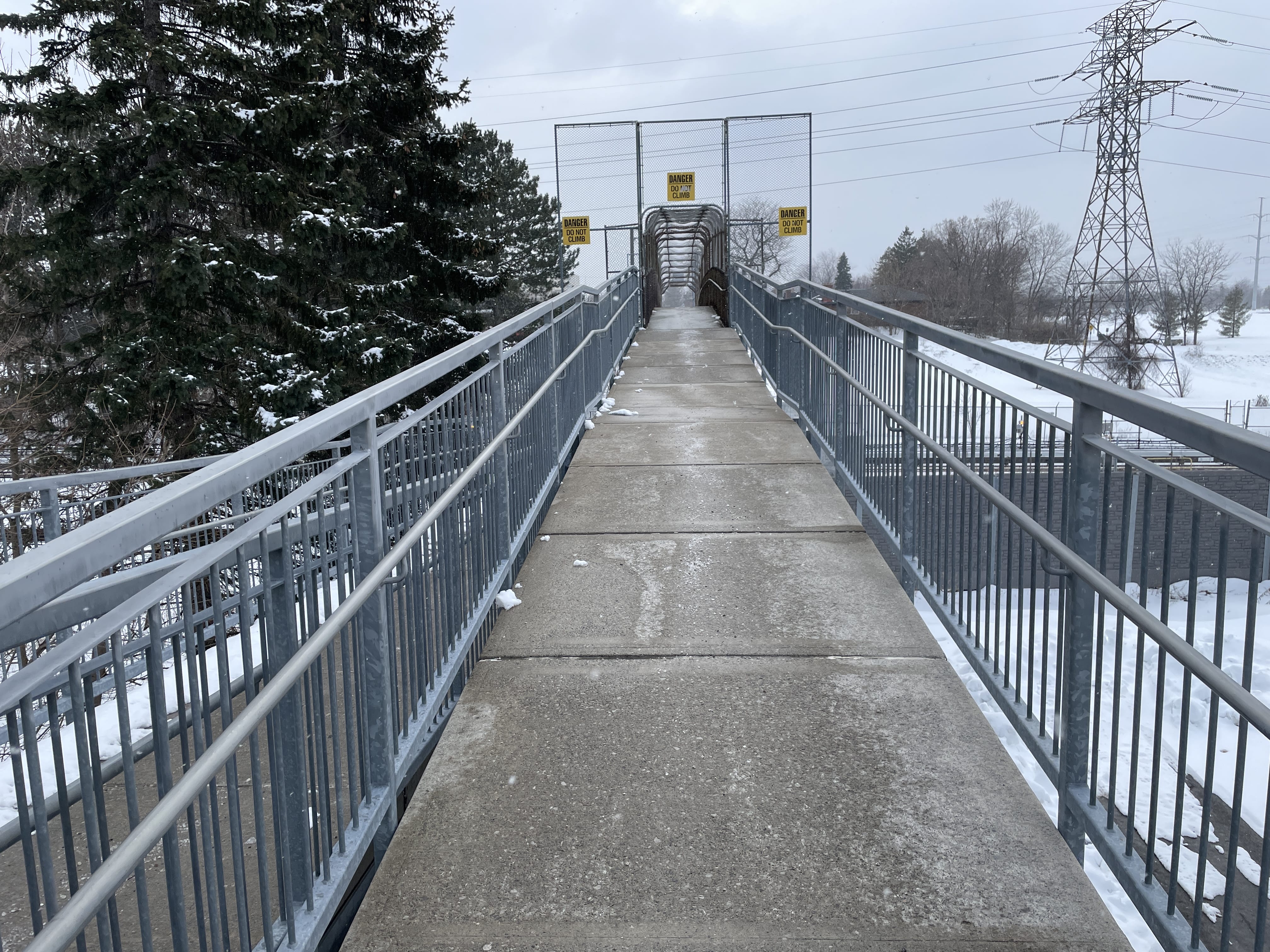 Daytime photo of the east side of the bridge over the rail line, which has straight ramps stacked back and forth down to ground level. The pavement is clear and visibly salted. In the background are some large evergreen trees and a power line.
